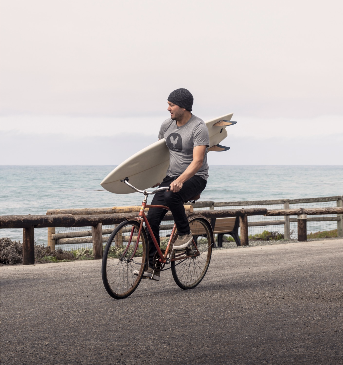 Man riding a bicycle with a surfboard on his back near a coastal road.