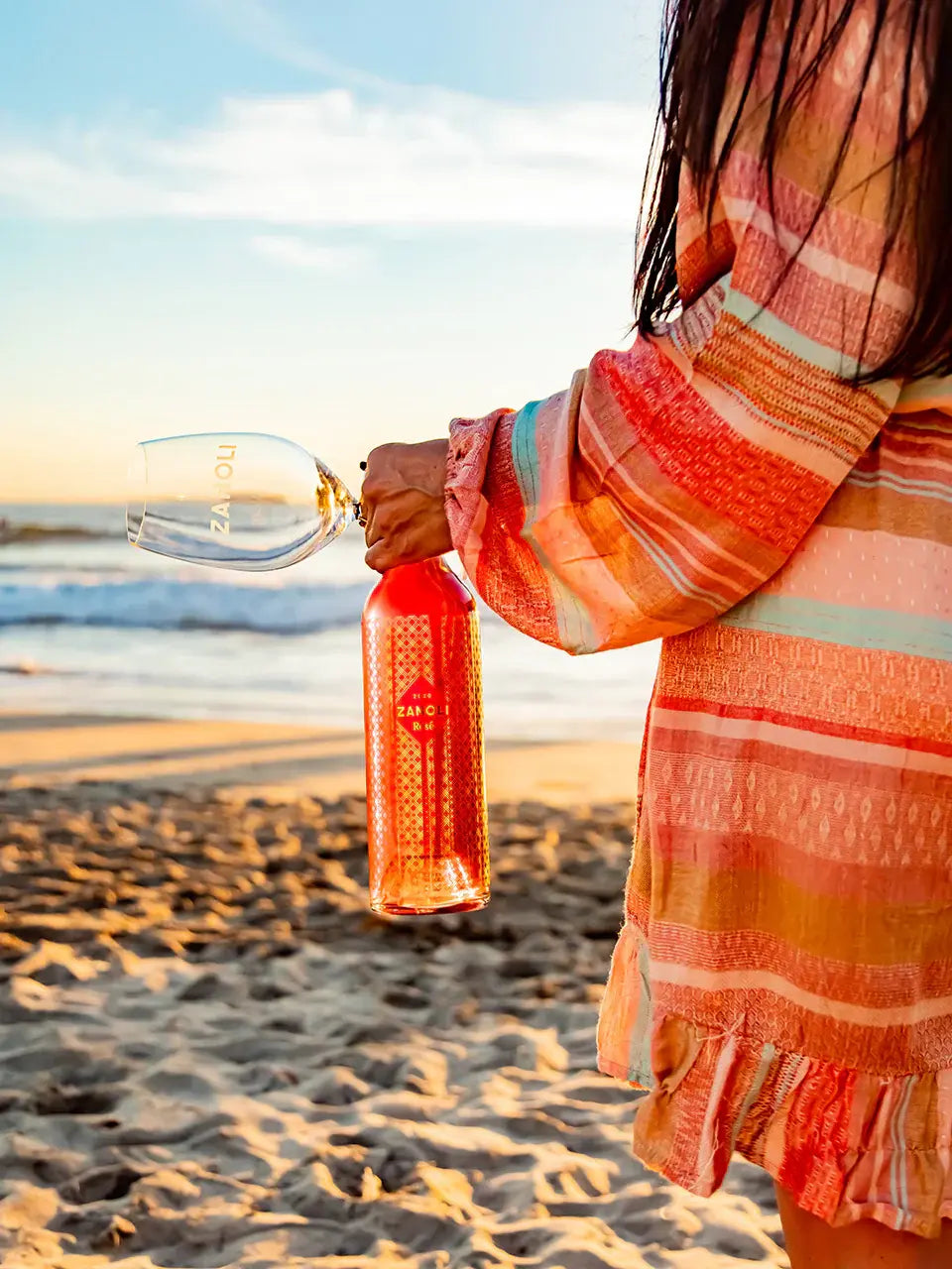 Person holding a red bottle with a pattern on a beach at sunset