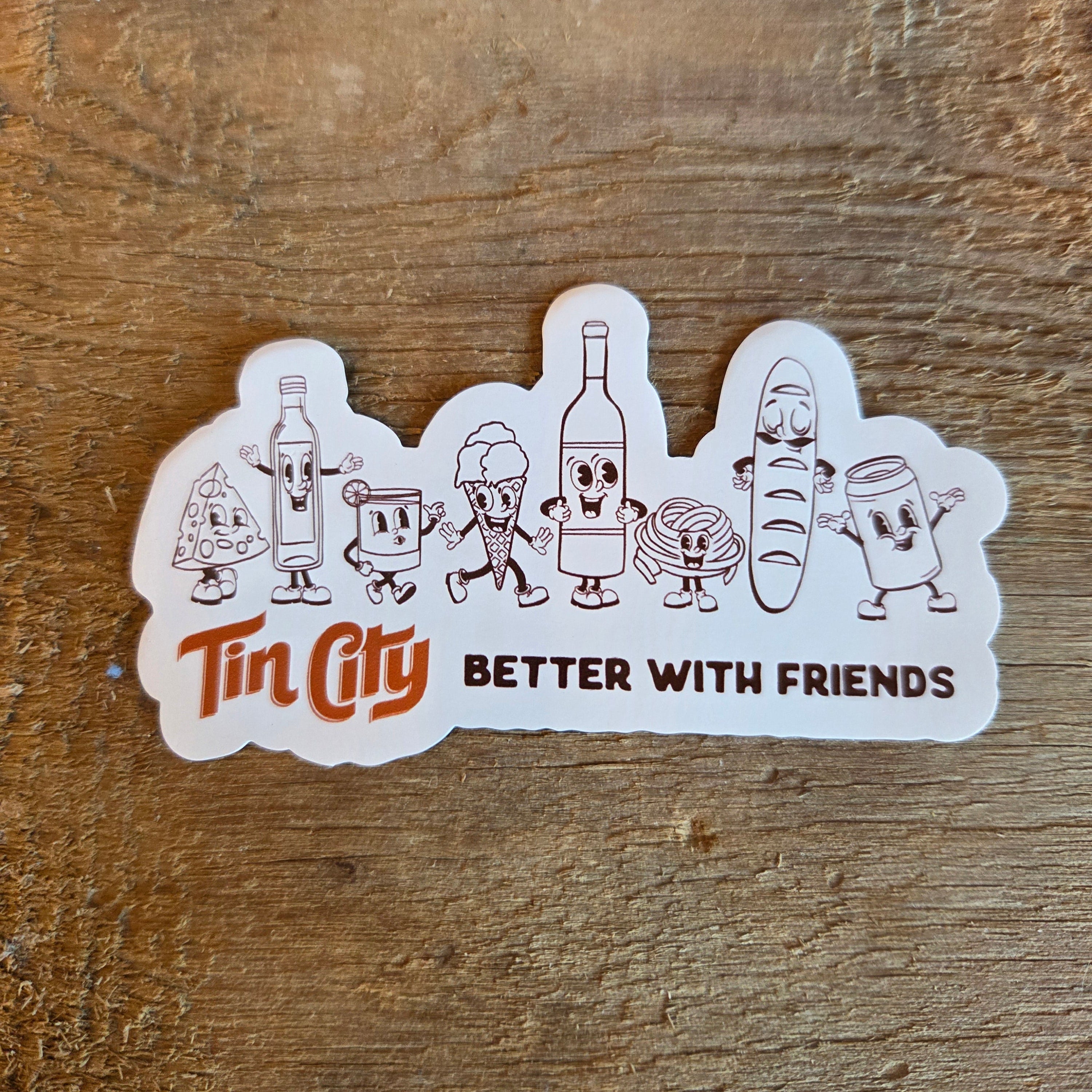 Magnet with cartoon characters and 'Tin City Better with Friends' text on a wooden surface